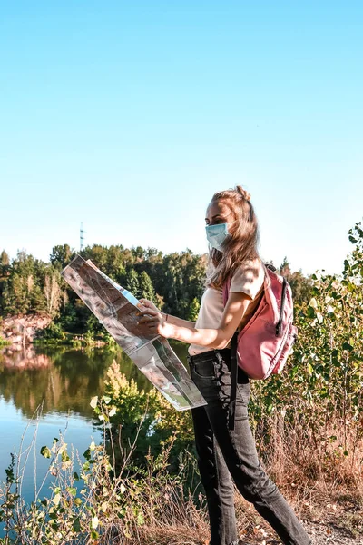 Young traveler wearing protective face mask and holding a map. Safe trip. Social distancing. Girl wearing backpack. Local travel. Beauty of nature