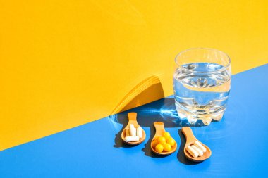 Bir bardak su ve ahşap kaşıkta haplar mavi ve sarı arka planda sert ışık ve gölgelerle. Modern izometrik yaratıcı minimalizm kavramı. Vitaminler, prebiyotikler, probiyotikler. Sonbahar vitamin dozu.