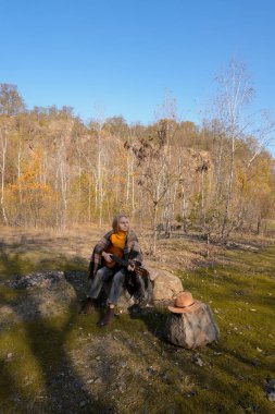 Güzel sarışın kız akustik gitar çalıyor ve şarkı söylüyor güneşli bir sonbahar gününde dışarıda eğleniyor. Hippi kadın boho stilinde öğreniyor ve müzik enstrümanı çalıyor. Sonbahar yaprakları