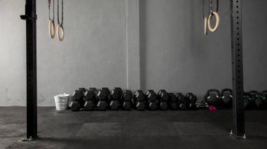 Dambıllar de crossfit de varios pesos con fondo gris sobre piso de goma de un Box con aros de gimnasia a los lados