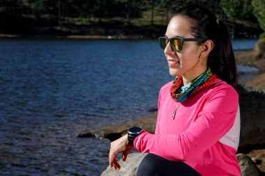 Koşucu kadın koşudan sonra akıllı saatini kullanıyor, göl kıyısında taş üstünde oturuyor.