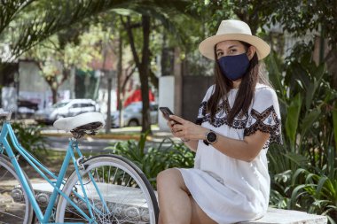 Yüzü maskeli kız cep telefonunu kontrol ediyor, parkta oturuyor, üzerinde elbise ve şapkası ve yanında da bisikleti var.