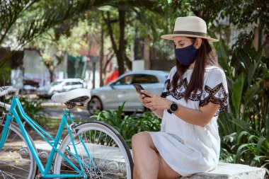 Yüzü maskeli kız cep telefonunu kontrol ediyor, parkta oturuyor, üzerinde elbise ve şapkası ve yanında da bisikleti var.