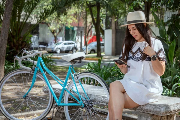 young latina woman checking her cell phone sitting in a park with a blue bicycle next to her