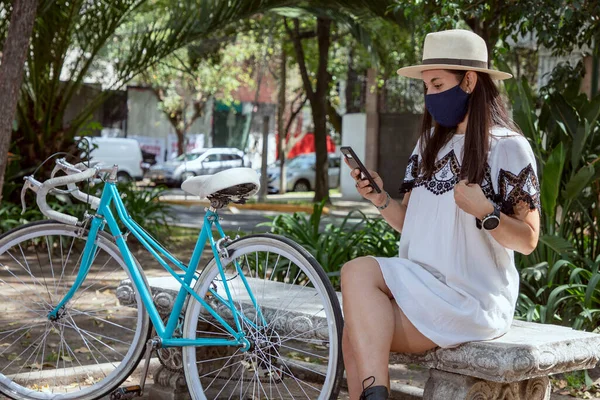 Girl with face mask checking her cell phone, sitting in the park wearing dress and hat with a bicycle by the side