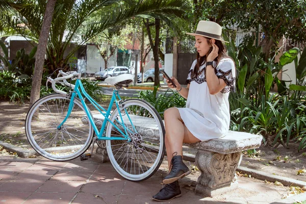 Genç Latin bir kadın cep telefonunu kontrol ediyor. Yanında mavi bir bisikletle parkta oturuyor.