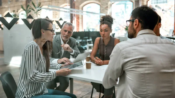 Using digital technologies. Young business woman holding digital tablet and discussing something with colleagues while sitting at the office table together