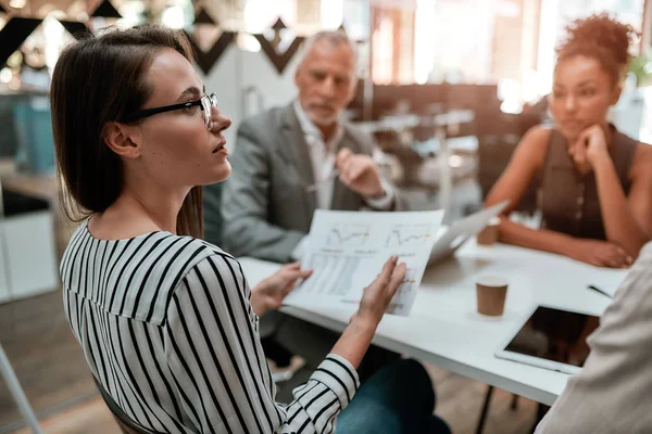 Analizando resultados. Joven mujer de negocios sosteniendo informe financiero y discutiendo algo con sus colegas mientras están sentados en la mesa de la oficina juntos — Foto de Stock