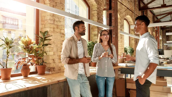 Joli discours. Trois jeunes collègues en tenue décontractée tenant des tasses à café et discutant de quelque chose tout en restant debout dans le bureau moderne — Photo