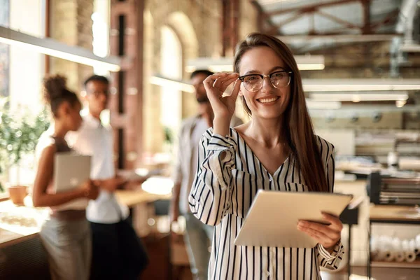 Wanita bisnis yang sukses. Wanita muda ceria memegang tablet digital dan melihat kamera dengan senyum sambil berdiri di kantor modern — Stok Foto