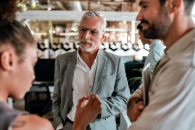Personel toplantısı. Resmi giyinmiş yakışıklı bir adam ofiste dururken genç meslektaşlarıyla bir şeyler tartışıyor.