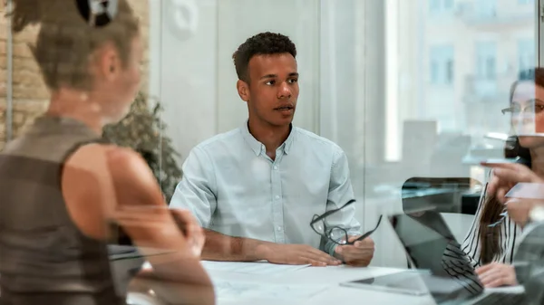 Experto en negocios. Joven afroamericano sosteniendo anteojos y explicando algo a sus colegas mientras está sentado en la mesa de la oficina detrás de la pared de vidrio en la oficina moderna —  Fotos de Stock
