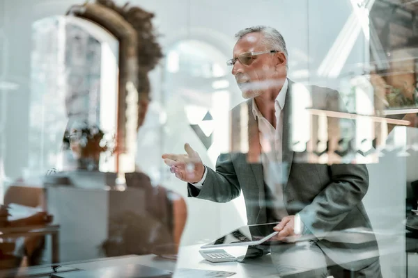 Brainstorming. Homme mûr sérieux en tenue formelle expliquant quelque chose à ses collègues et gesticulant tout en étant assis à la table du bureau derrière le mur de verre dans le bureau moderne — Photo