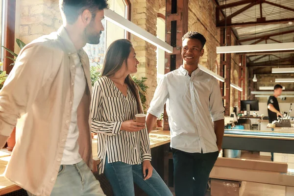 Pauzeer op het werk. Jonge en vrolijke collega 's in casual dragen praten over iets en houden koffiebekers terwijl ze in het moderne kantoor staan — Stockfoto