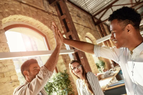 Erfolge feiern. zwei gut gelaunte Männer geben High-Five, während sie mit Kollegen im modernen Büro stehen — Stockfoto