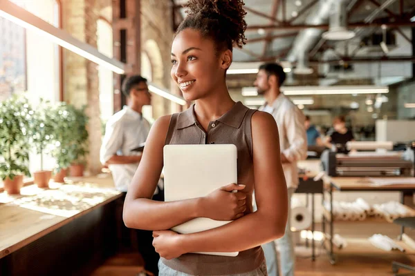 Wanita bisnis modern. Wanita muda dan ceria afro american memegang laptop dan tersenyum sambil berdiri di kantor modern — Stok Foto