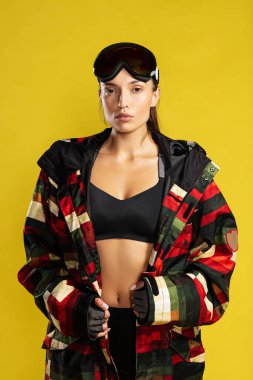 Kendine güvenen bir kayakçı. Stüdyoda sarı arka planda dikilirken kameraya bakan kayak kıyafeti giyen güzel bir kadın. Kayak