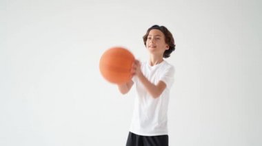 Basketbol hayranıyım. Aktif bir gencin portresi beyaz tişörtlü basketbol topu fırlatırken gri arka planda kameraya gülümseyen mutlu çocuk.