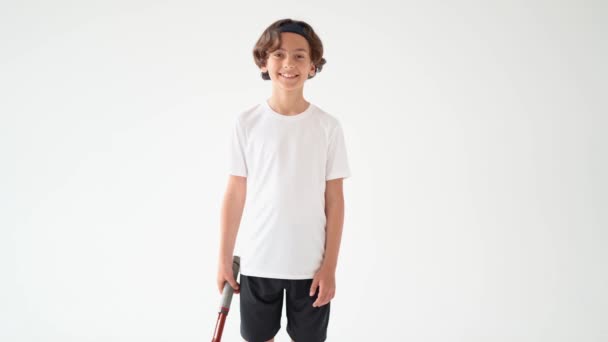 นักเทนนิสตัวน้อย วัยรุ่นชายที่มีความสุขในเสื้อยืดสีขาวถือไม้เทนนิสและมองไปที่กล้องด้วยรอยยิ้มแยกกันบนพื้นหลังสีขาว — วีดีโอสต็อก