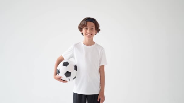 Jugador de fútbol. Retrato de un lindo niño feliz en camiseta blanca sosteniendo la pelota de fútbol en una mano y sonriendo a la cámara mientras está de pie sobre un fondo gris — Vídeo de stock