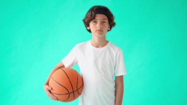 Çocuklar için basketbol. Beyaz tişörtlü, elinde basketbol topu olan, kameraya bakıp gülümseyen, turkuaz mavi arka planda duran aktif bir gencin portresi.