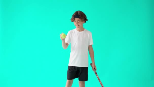 เล่นเทนนิส เด็กวัยรุ่นที่มีความสุขในเสื้อยืดสีขาวถือไม้เทนนิสและลูกเทนนิส ยิ้มที่กล้อง ยืนต่อต้านพื้นหลังสีน้ําเงินเทอร์ควอยส์ — วีดีโอสต็อก