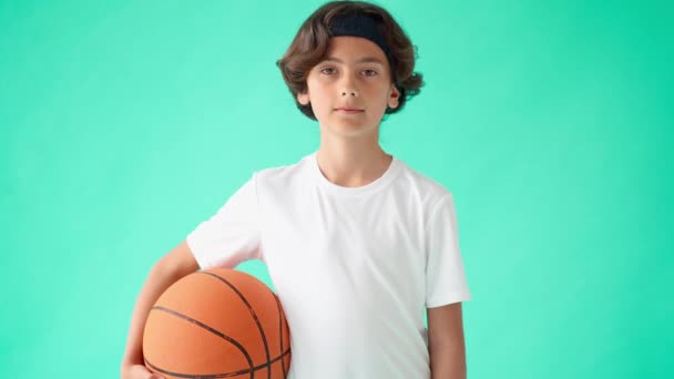 Молодий баскетболіст. Портрет активного хлопчика-підлітка в білій футболці, що тримає баскетбол і посміхається на камеру, стоїть на тлі бірюзового синього фону — стокове відео