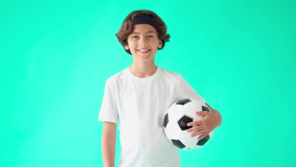 Menino adolescente feliz em camiseta branca segurando bola de futebol em uma mão e rindo da câmera enquanto estava de pé contra o fundo gradiente verde — Vídeo de Stock