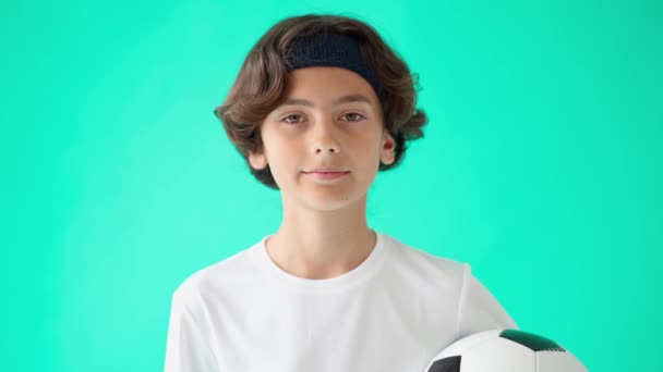 Портрет милого подростка в белой футболке, держащего футбольный мяч, смотрящего в камеру и смеющегося, стоящего на бирюзовом синем фоне — стоковое видео