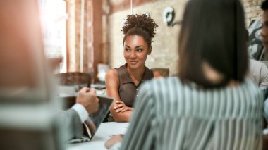 Burada çalışmaktan mutluyum. Genç ve neşeli Afro-Amerikan kadın, modern ofisteki meslektaşlarıyla görüşürken gülümsüyor ve yan bakıyor.