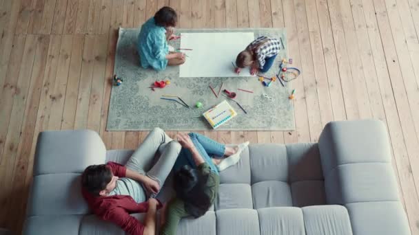 Kreatív tevékenység. Top kilátás két kisgyerek, testvér és testvér rajz ceruzával, feküdt a földön, míg a fiatal szülők pihentető a kanapén — Stock videók