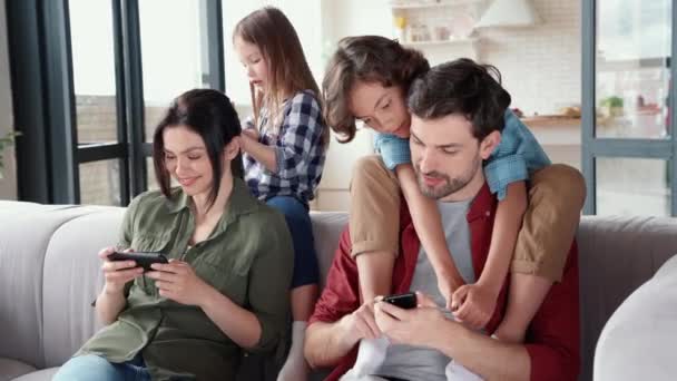 주말을 즐기는 거야. 네 식구로 된 행복 한 가족 이 거실의 소파에서 함께 휴식을 취하고 있다. 어린 부모들은 스마트폰을 사용하고 웃으면서 귀여운 어린 아이들 과 집에서 즐거운 시간을 보내고 있습니다 — 비디오