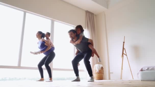 Spor ailesi evde birlikte spor yapıyor. İki heyecanlı çocuğu sırtında taşıyan genç ve güzel ebeveynler evdeki parlak modern odada çömelme hareketleri yapıyorlar. — Stok video