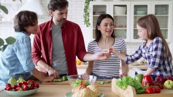 Χαρούμενη νεαρή οικογένεια που μαγειρεύουν μαζί στη σύγχρονη κουζίνα στο σπίτι, μιλούν και επαινούν ενώ ετοιμάζουν το φαγητό. Μητέρα και πατέρας διδάσκουν σε δύο μικρά παιδιά πώς να κόβουν φρέσκα λαχανικά — Αρχείο Βίντεο