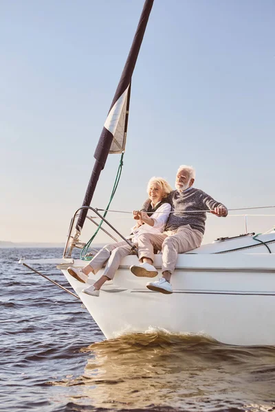 A happy senior couple talking, sitting on the side of a sail boat on a calm blue sea. Man hugging his woman while enjoying view — Stock Photo, Image