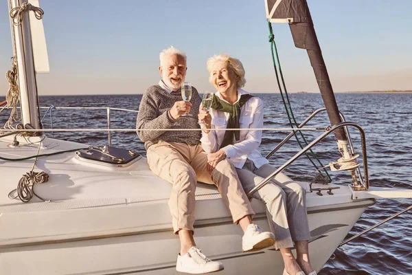 Denizde yüzen yelkenli ya da yat güvertesinde oturan mutlu yaşlı çift. Kadın ve erkek şarap içip gülüyorlar.