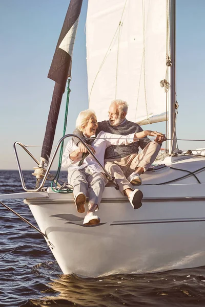 航行是一种乐趣。一对快乐的老夫妇坐在平静的蓝色海面上的帆船边。男人在和女人说话的时候拥抱她 — 图库照片