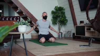 Evde kal. Siyah koruyucu maskeli güçlü atletik adam evde yoga minderinde oturuyor, ısınıyor ve egzersiz yapıyor, dizüstü bilgisayarda online dersler izliyor.