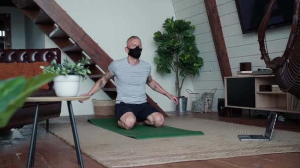 Blijf thuis. Sterke atletische man draagt zwart beschermend masker zittend op yoga mat thuis, opwarmen en sporten, kijken naar online klasse op laptop — Stockvideo