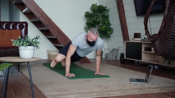 Entrenamiento en casa. Hombre atlético fuerte con máscara protectora negra y ropa deportiva haciendo ejercicio en casa, viendo clases en línea en el portátil — Vídeos de Stock