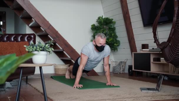 Thuis training online. Sterke atletische man draagt beschermende masker doen push-ups op yoga mat tijdens het sporten thuis, kijken naar online klasse op laptop — Stockvideo