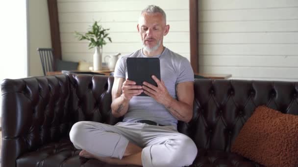 Colloquio. Bello uomo maturo seduto sul couh a casa, utilizzando tablet digitale e parlando online tramite video chat. Comunicazione a distanza — Video Stock