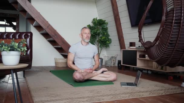 Практикує йогу онлайн. Розслаблений зрілий чоловік, сидячи в лотосі позує на йога килимок у вітальні і виконує дихальні вправи, роздумуючи вдома — стокове відео