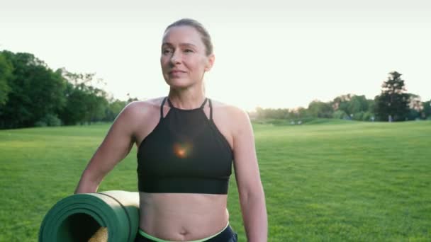 Mooie gelukkige vrouw in sportkleding met yoga mat, wandelen over een groen mooi veld op een zomerochtend en genieten van het geweldige uitzicht — Stockvideo