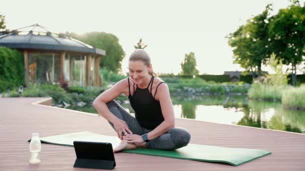 Yoga online onderwijzen. Gelukkig trekken vrouw in sportkleding kijken naar digitale tablet en het geven van online yoga klasse terwijl zitten op een mat in het park op een zomerochtend — Stockvideo