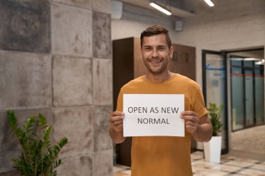 Genç, mutlu bir adam, ofis çalışanı, kameraya yeni noel olarak kağıt gösteriyor ve modern çalışma alanında dururken gülümsüyor.