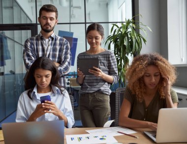 Modern ofiste birlikte çalışan bir grup genç çok kültürlü iş adamı, dizüstü bilgisayar, tablet, akıllı telefon kullanıyorlar. Ortak çalışma alanında başarılı proje ekibi