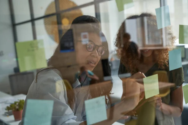 Brainstorming. Jeune femme métisse portant des lunettes écrivant quelque chose sur des notes collantes sur un panneau de verre tout en travaillant avec des collègues dans le bureau moderne — Photo
