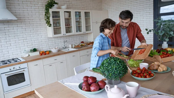 Sharing skills. Young dad and his little happy son cutting preparing a vegetarian salad together in the modern kitchen at home, spending time together