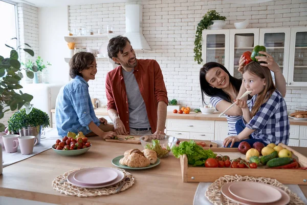 Preciosa familia con niños felices divirtiéndose mientras cocinan juntos en la cocina moderna en casa, preparando una ensalada y jugando con pimiento dulce — Foto de Stock